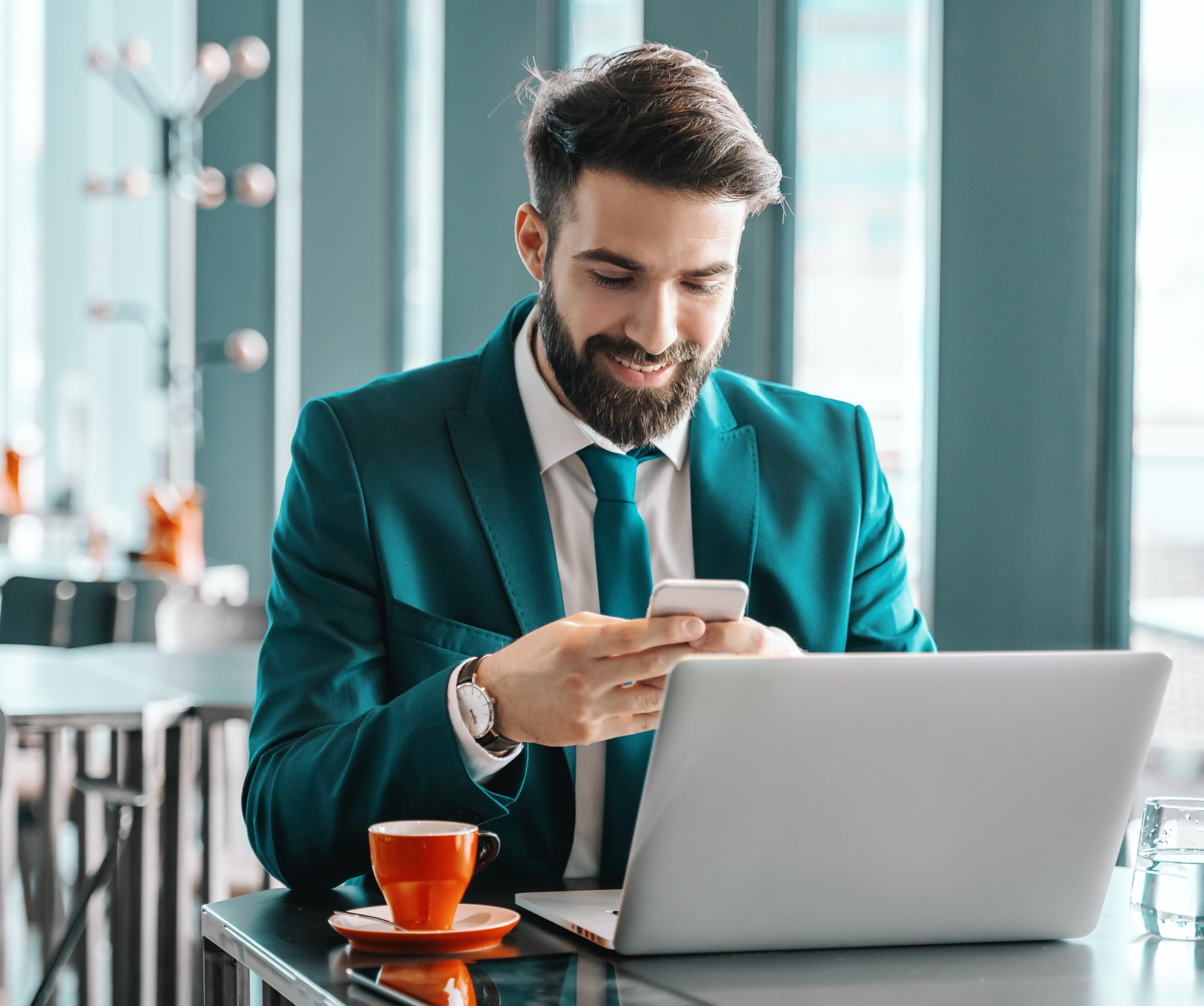 Smiling attractive ambitious businessman in turquoise suit using smart phone for reading or writing message while sitting in cafe.